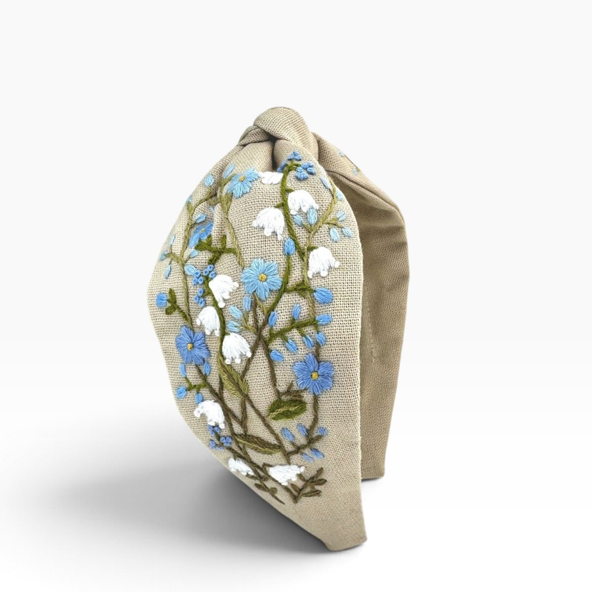 A beige fabric headband with an embroidered floral pattern of blue forget-me-nots and white blossoms on a light gray background.