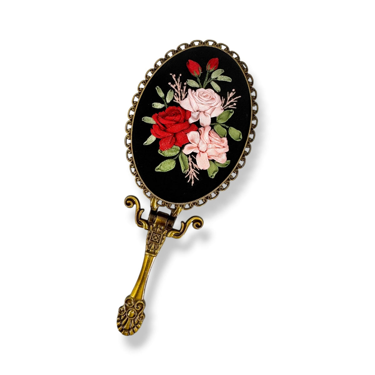 An ornate oval hand mirror with a black face featuring embroidered red and pink roses with green leaves, set in an antique gold-toned metal frame and handle.