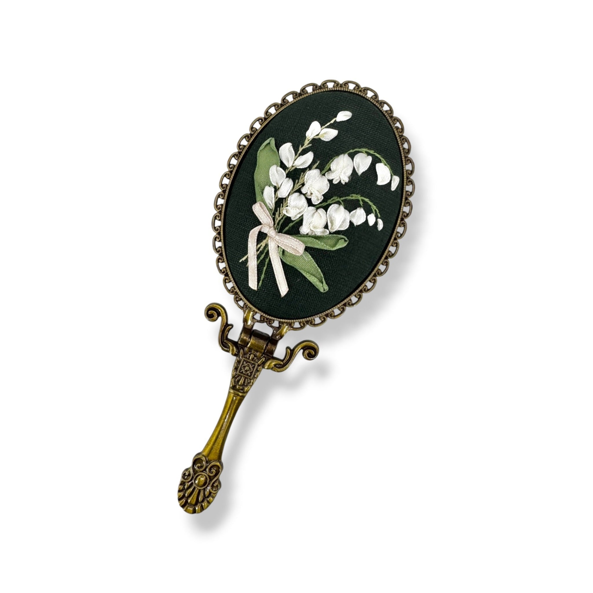 An antique-style oval hand mirror with a dark green face depicting white lily of the valley flowers and green leaves, set in an ornate antique bronze metal frame with a decorative handle.