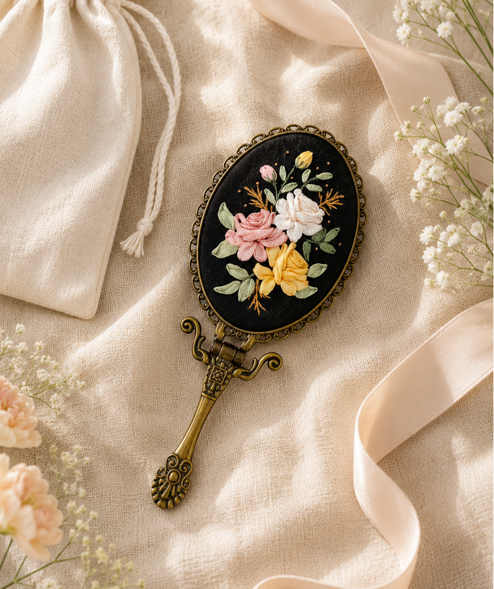 An oval hand mirror with a black background and colorful floral embroidery, featuring an ornate antique bronze handle, styled on a beige linen fabric with a cream drawstring pouch, white baby's breath, and a pale pink ribbon.